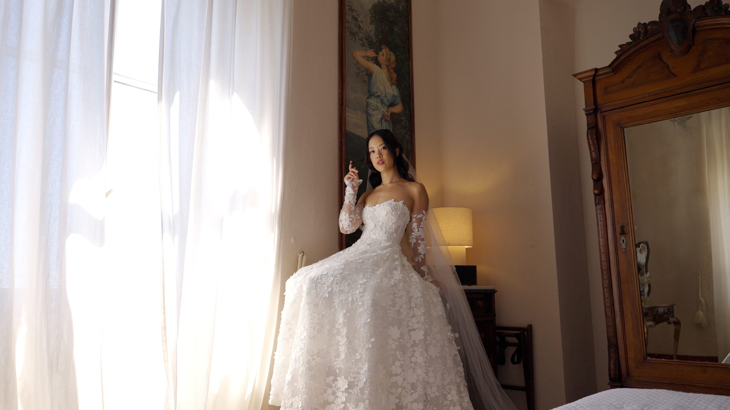 Gaia standing beside a bedroom window, showing the full skirt of her lace wedding gown.