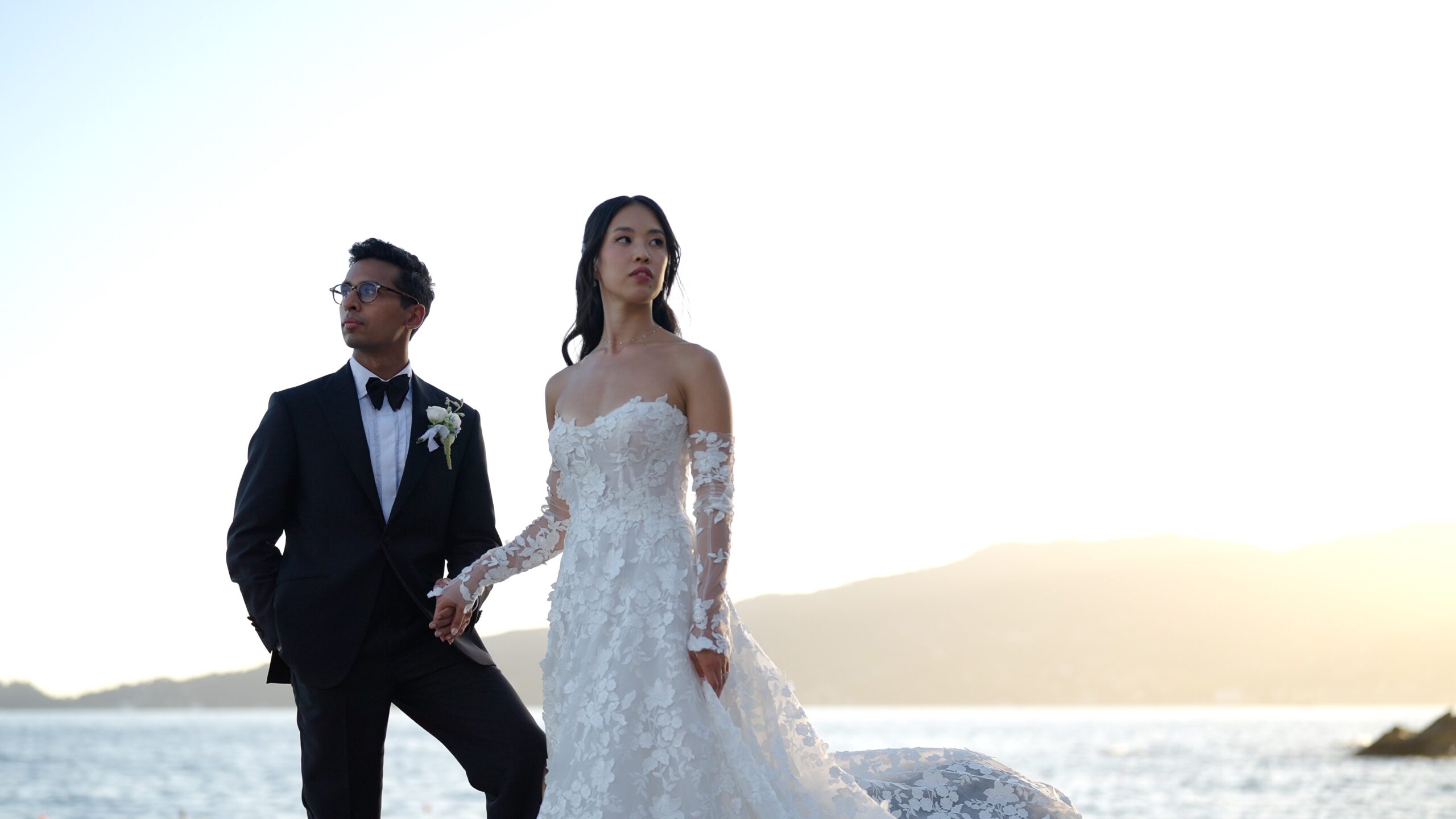 Gaia and Sheh holding hands on seaside rocks at sunset.