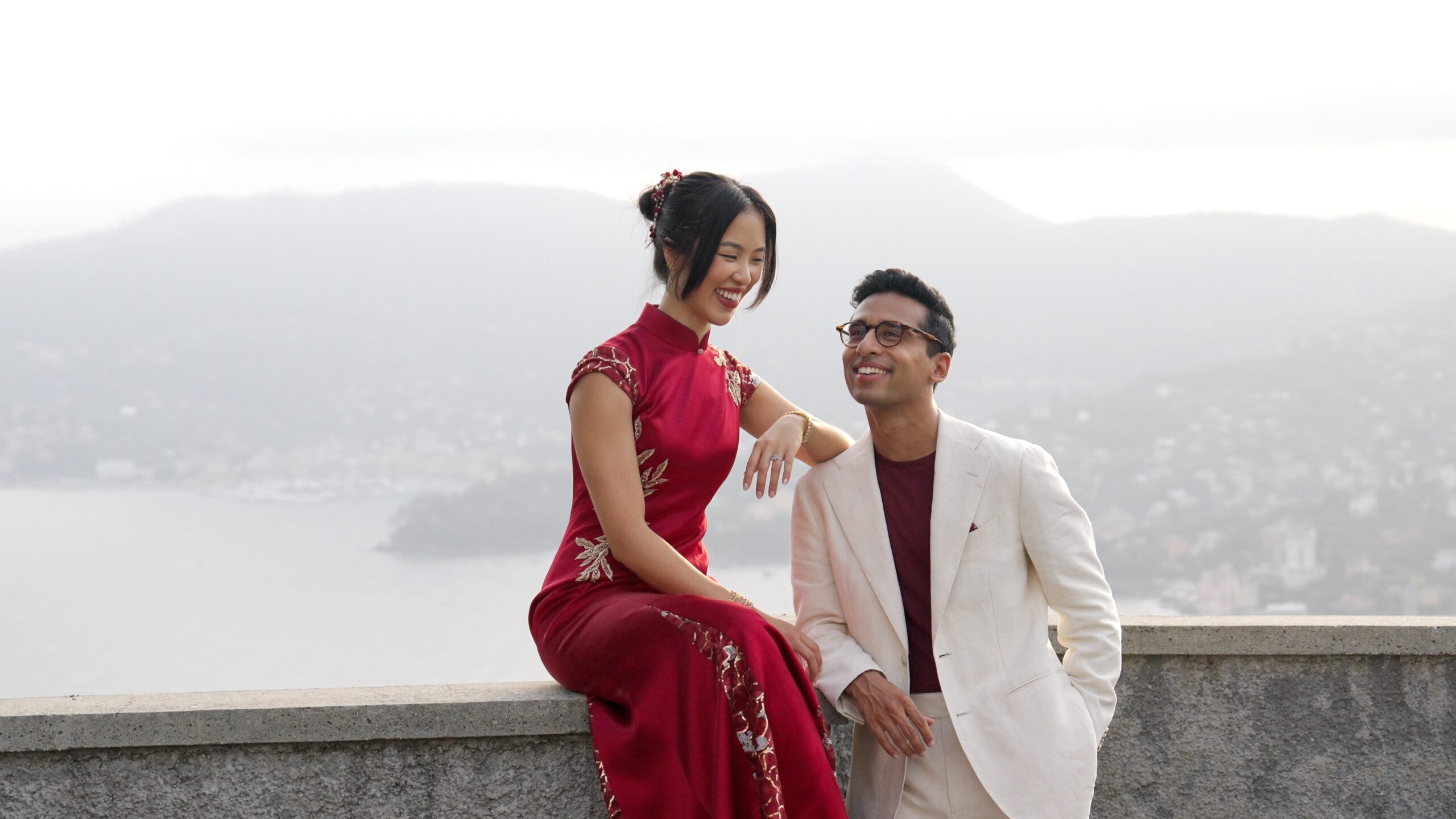 Gaia in a red dress and Sheh in a white jacket sitting on a terrace overlooking the coast.