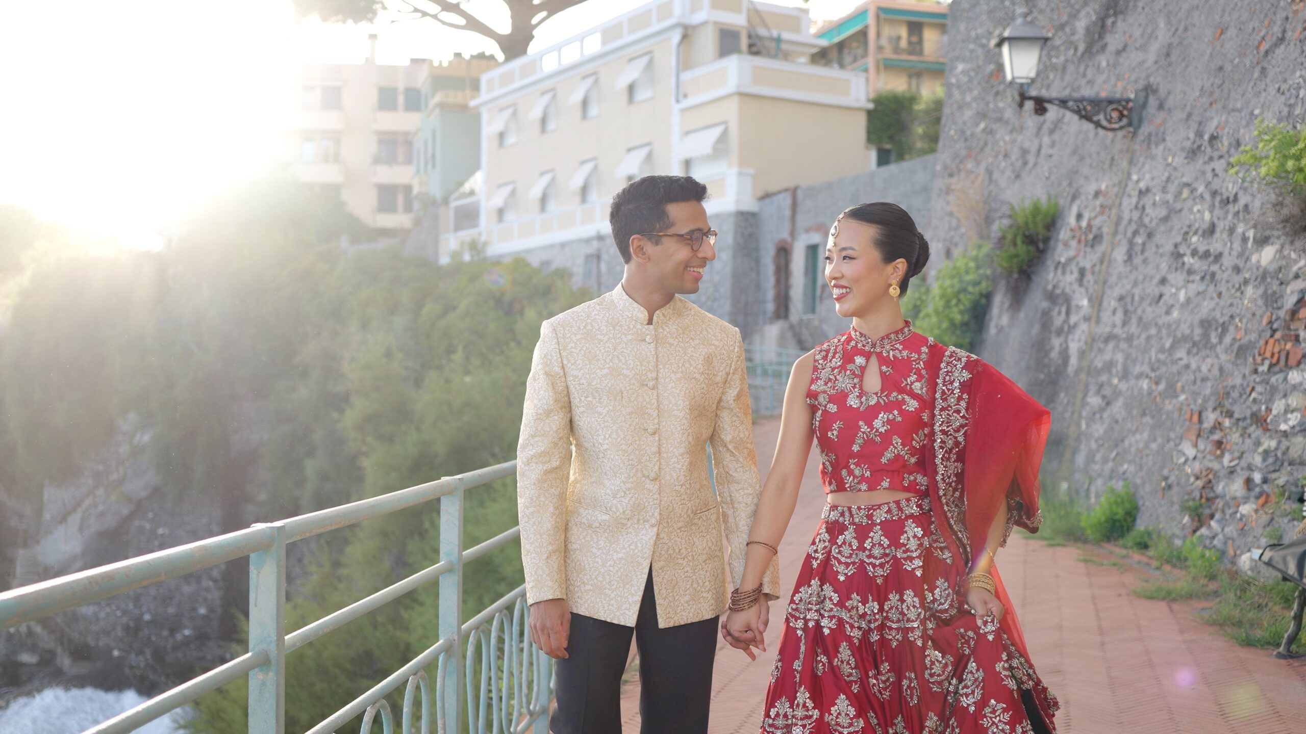 Gaia in a red embroidered outfit and Sheh in an ivory jacket walking along a seaside path.