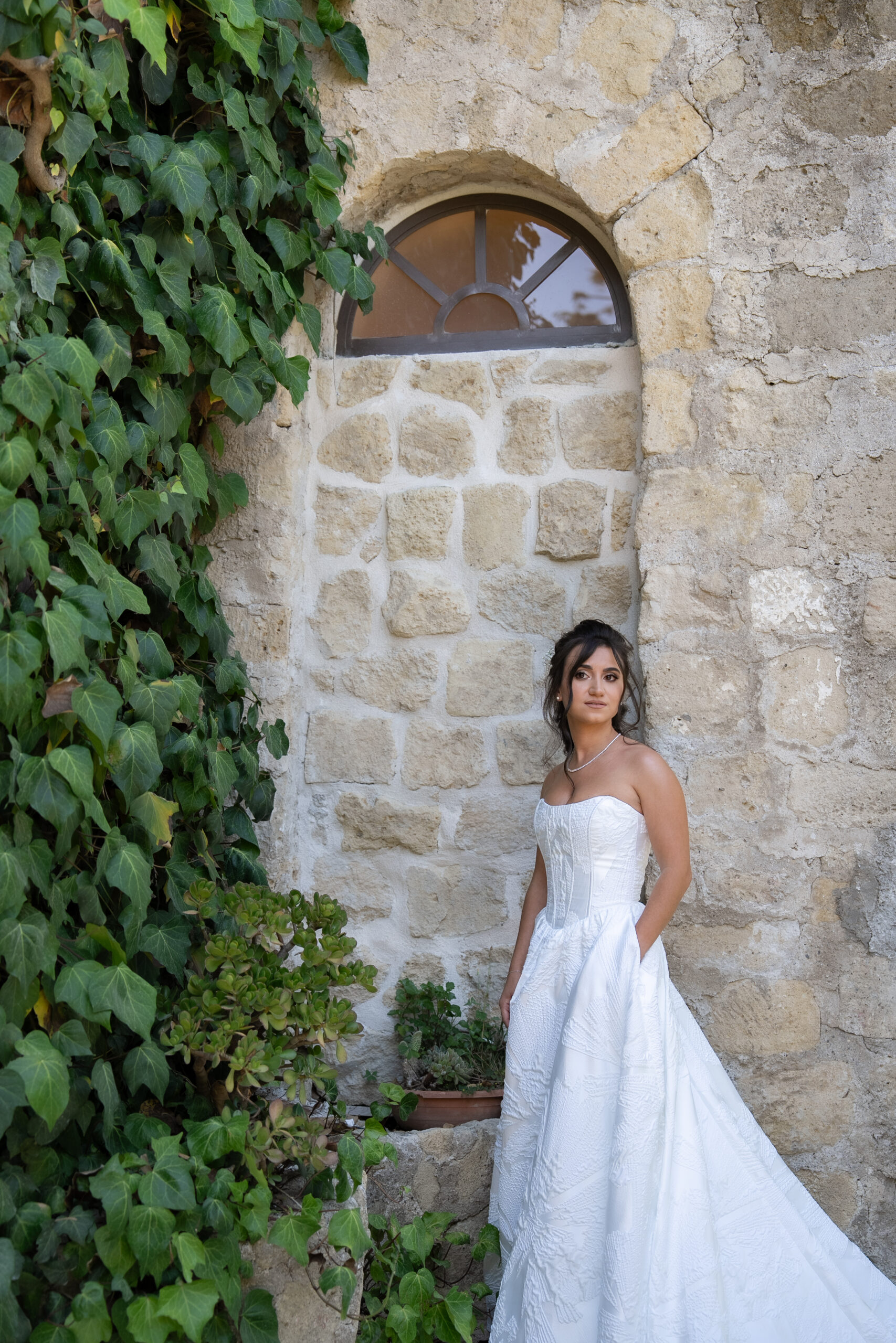 Monica in her wedding dress beside an ivy covered stone wall and an arched window.
