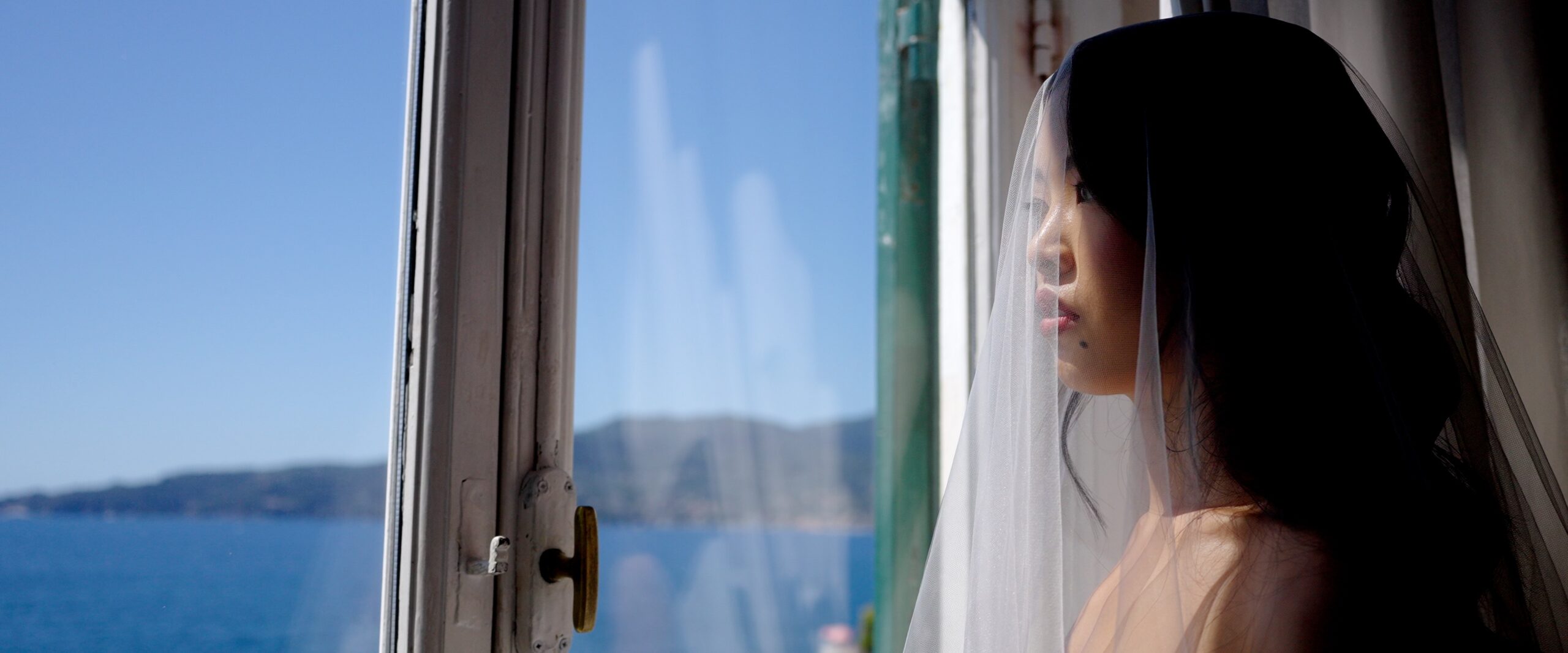 Gaia looking through a window towards the sea during her bridal preparations.